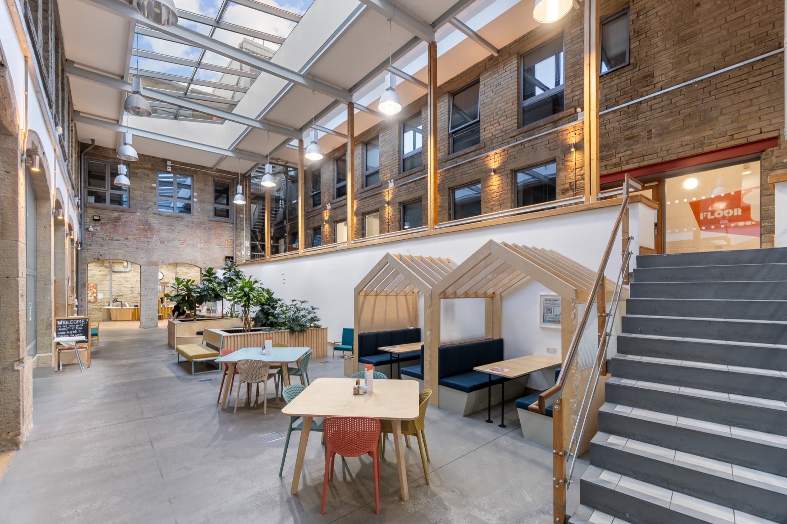 Atrium beneath a glazed roof, with exposed brick and stone, timber booth seating shaped like small huts, cafe tables and a staircase rising to the first floor.