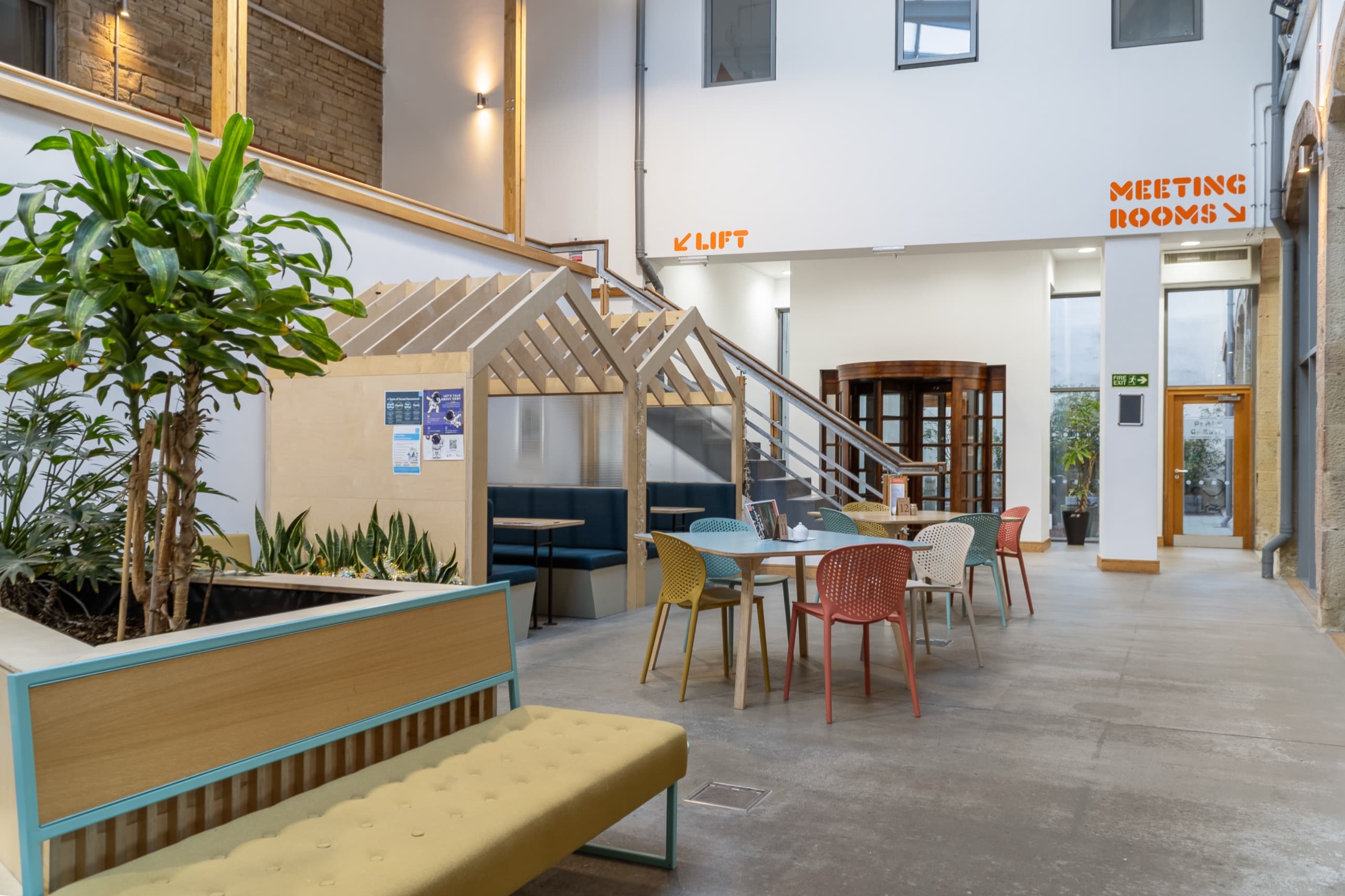 Timber booth seating and cafe tables in the atrium, with orange lettering on the wall above pointing to the lift and the meeting rooms.