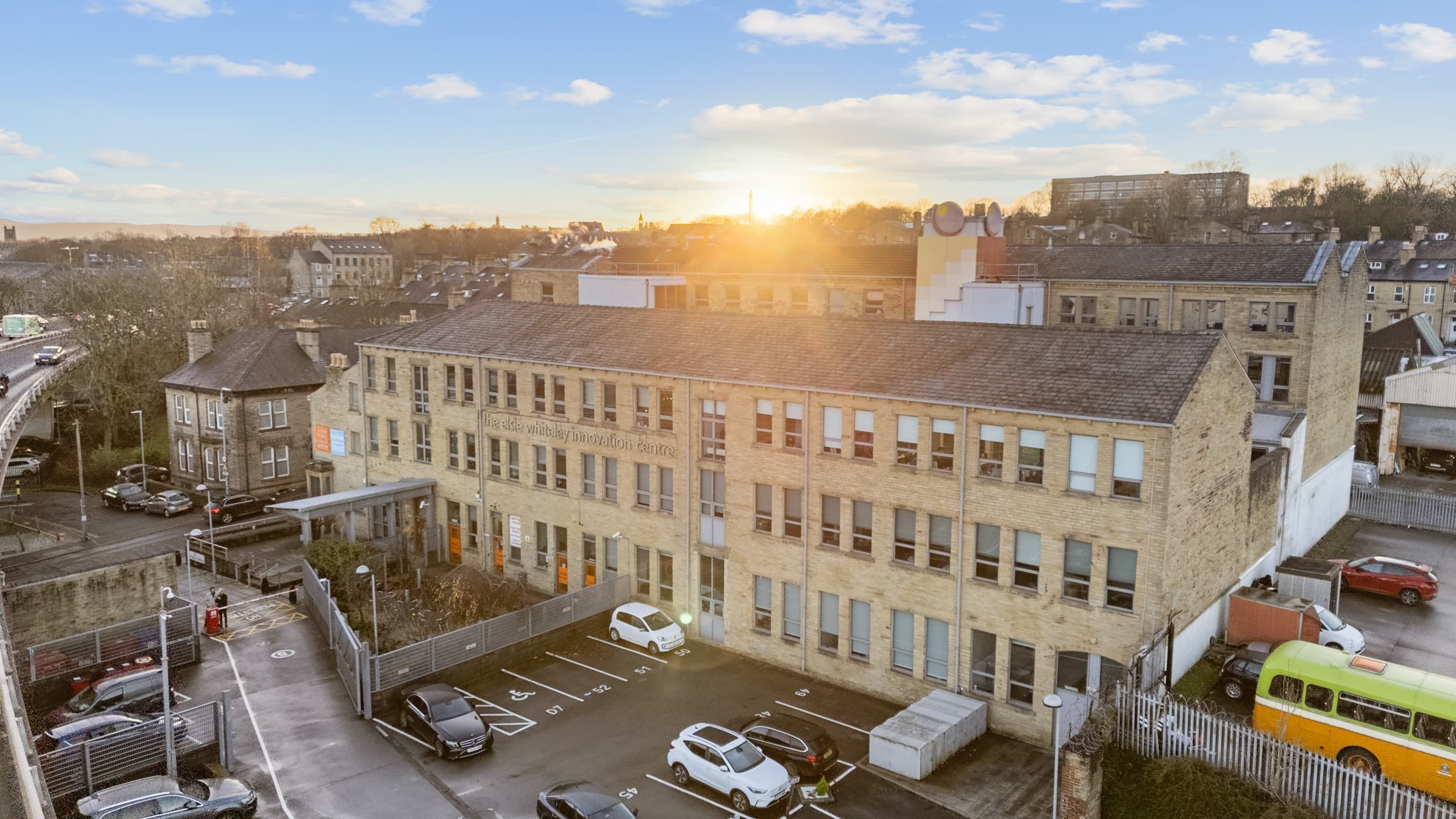 Aerial view of the Elsie Whiteley Innovation Centre at sunset, a three-storey stone mill building with its name lettered across the facade and marked parking bays in front.