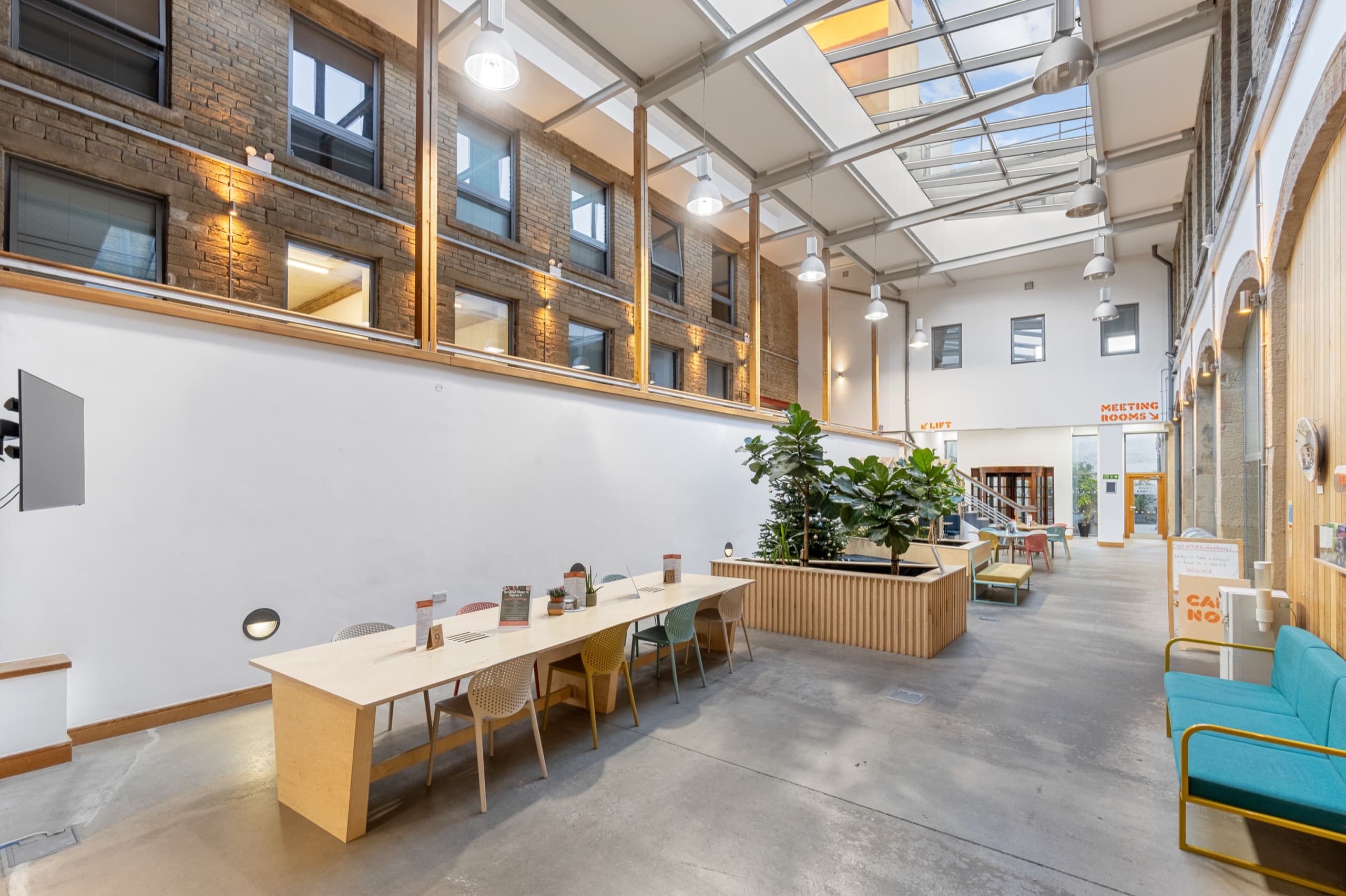Long shared work table with chairs running along the atrium wall, daylight falling through the glazed roof onto the polished concrete floor.