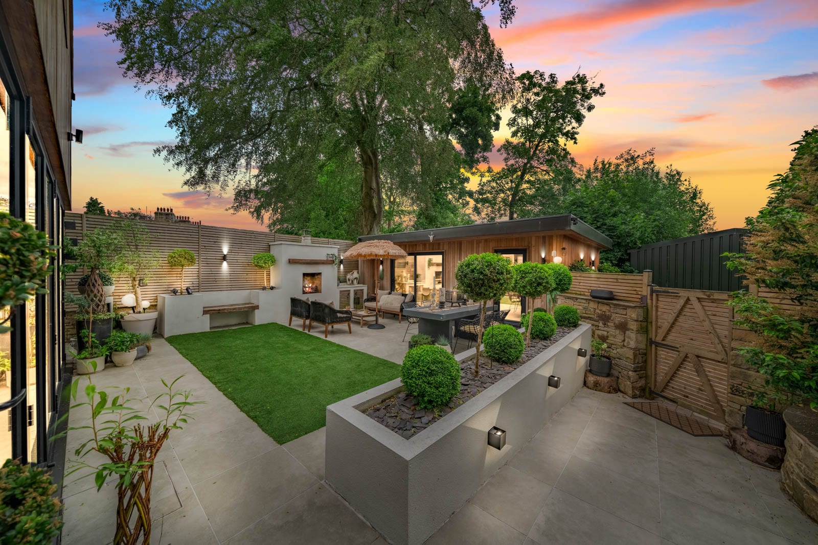 Landscaped walled garden at dusk with a cedar-clad garden room, an outdoor fireplace, topiary in a raised planter and a neat lawn under a pink evening sky.