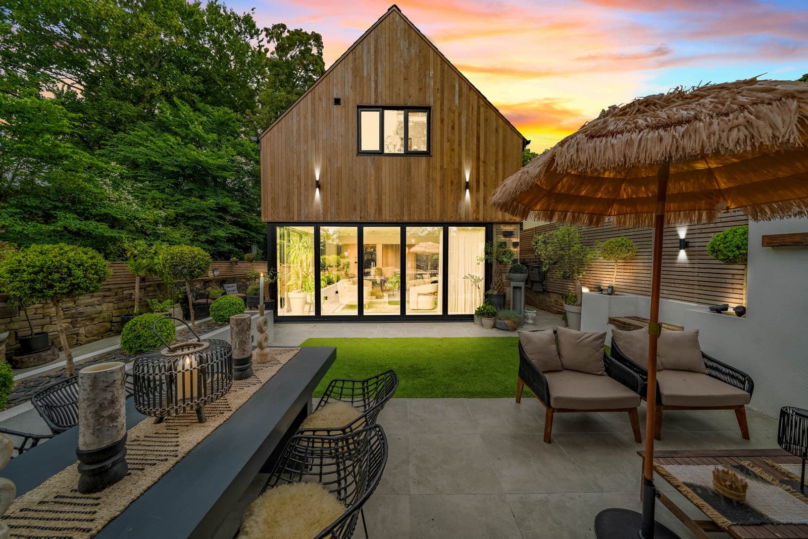 Timber-clad rear elevation lit at dusk, glazed doors glowing over a landscaped garden with a dining table and parasol.