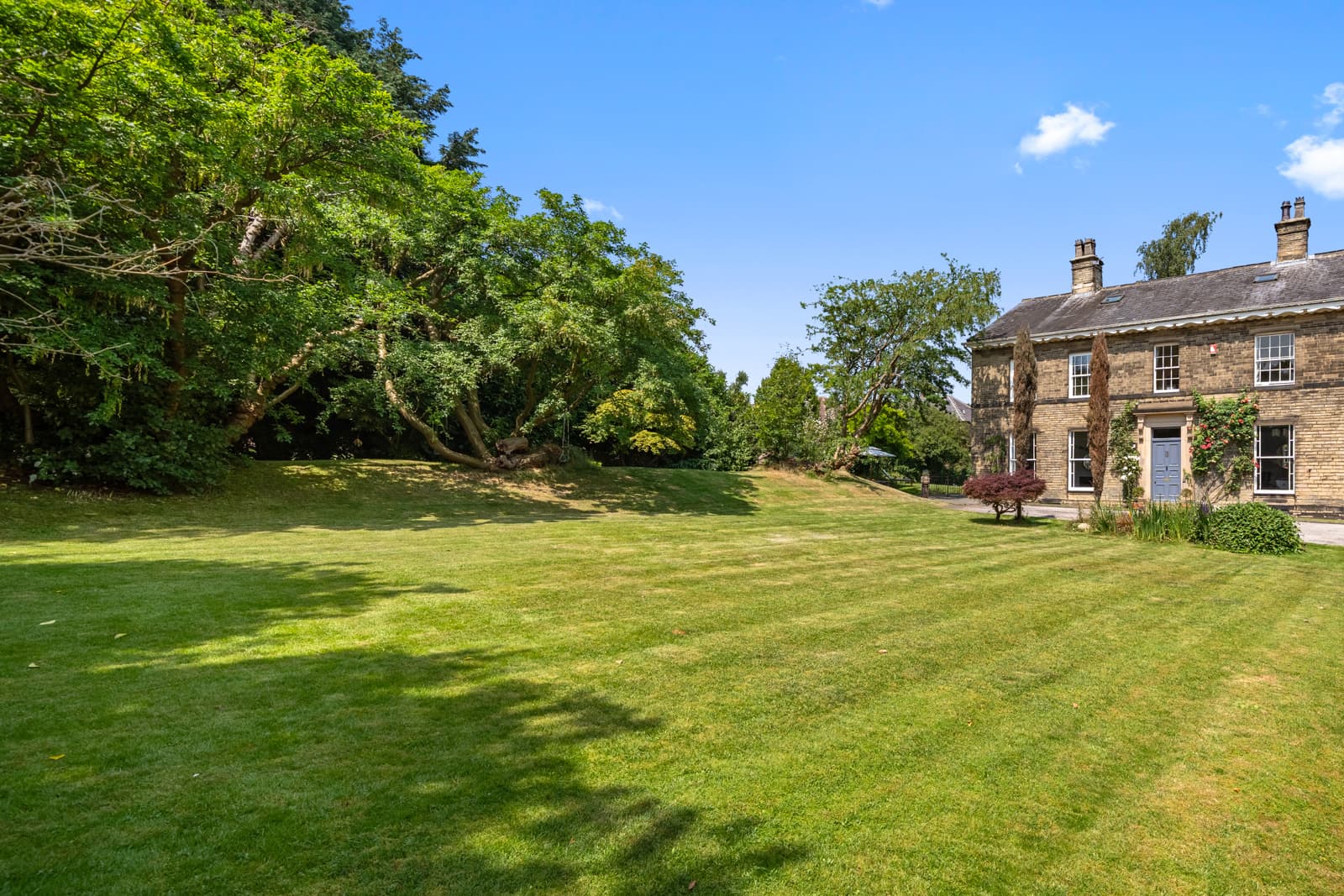 Stone period house at the edge of a broad striped lawn, mature trees along the boundary under a clear sky.
